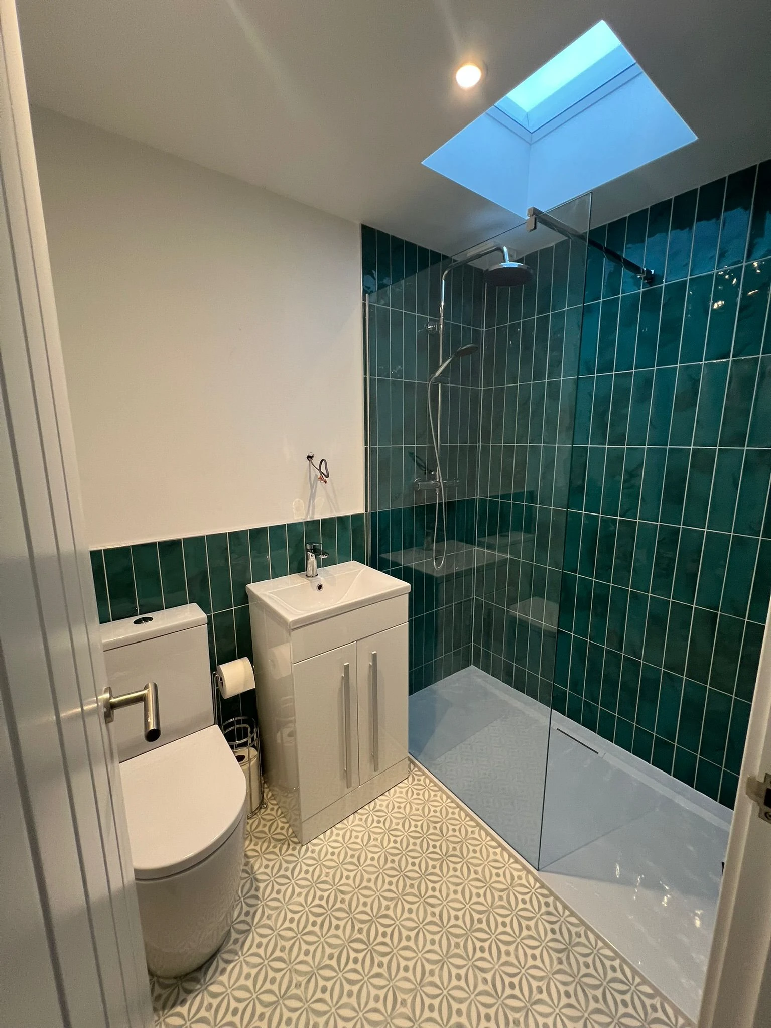 Modern bathroom with teal tiled walls, a glass shower, compact sink unit, toilet, and patterned floor tiles. Natural light from a skylight.